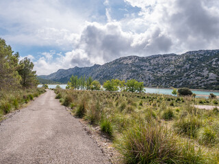 Fototapeta premium Gorg Blau artifical Lake, Mallorca, scenic dirt road winds through a grassy field, leading to a tranquil lake surrounded by rolling hills under a partly cloudy sky, Majorca