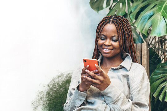 A cheerful woman with braided hair holds her smartphone in her hands surrounded by bright green nature