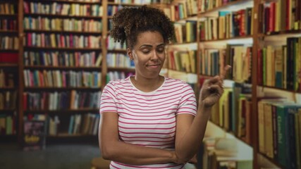 Woman pointing finger with crossed arms wearing striped shirt at books on wooden shelves in a building library aisle; skepticism.