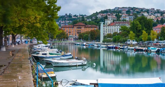 Rijeka, Croatia - August 21, 2025: Timelapse, zoom out transition. Quiet waterfront with small boats moored along a calm canal, reflecting colorful buildings, cloudy sky.