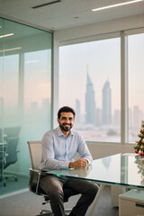 A smiling businessman sits at a glass desk in his modern office, overlooking the Dubai skyline through large windows.