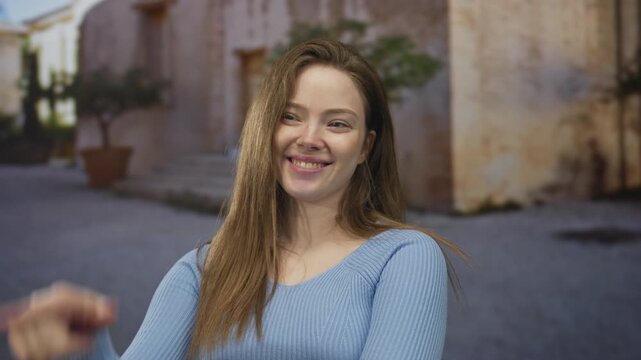 Woman with long hair and blue sweater smiling points finger to nose on sunlit old town cobblestone street; playful moment.