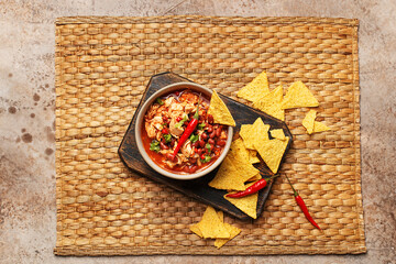 Traditional mexican dish chili con carne in ceramic bowl served on wooden board with chili peppers and nachos corn chips over woven placemat. Overhead view