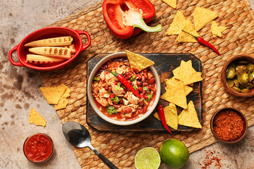 Traditional mexican dish chili con carne in ceramic bowl served on wooden board with nachos and various vegetables, sauces and spices over woven placemat. Overhead view, flat lay