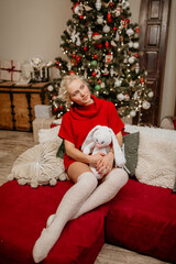 Obraz premium Happy blonde woman in a red sweater dress and cozy knee-high socks sitting on a couch, hugging a large knitted rabbit toy next to a brightly decorated Christmas tree