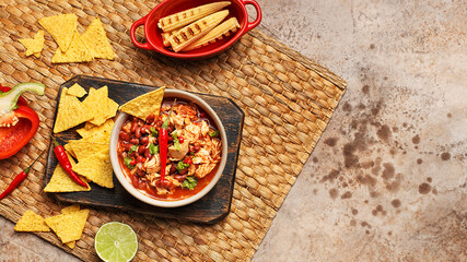 Traditional Mexican dish chili con carne in ceramic bowl served on wooden board with nachos, lime, chili pepper, bell pepper and grilled corn over beige concrete background. Top view, copy space