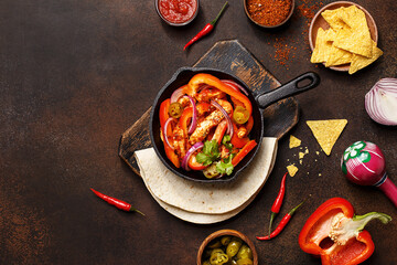 Traditional mexican dish fajitas in cast iron pan with tortilla, nachos, vegetables, sauces and maracas on dark concrete background. Flat lay, overhead view, copy space