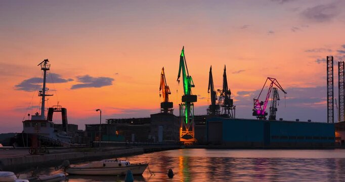 Pula, Croatia - August 17, 2025: Timelapse, zoom out transition. Industrial harbor at sunset with colorful cranes, docked boats, and soft pastel sky reflecting warm tones on the calm sea.