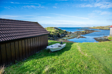 Fototapeta premium Serene coastal scene features a wooden shed and a small white boat on a grassy slope, with a clear blue sky and calm sea in the background, Haugesund, Norway