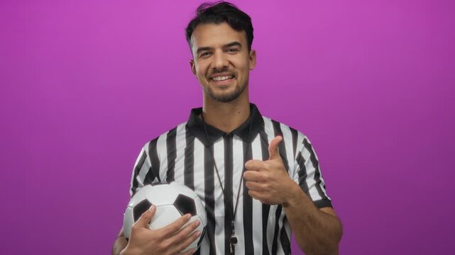 Young hispanic man wearing referee uniform holding soccer ball with thumbs up against isolated pink background, expressing positivity and sports enthusiasm.
