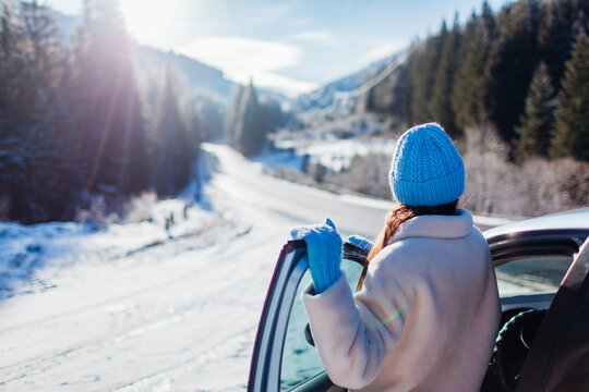 Back view of woman traveler admiring snowy mountains walking out of car looking at winter landscape. Traveling by auto