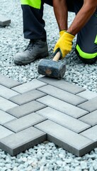 Worker laying gray pavers in herringbone pattern with mallet and gloves.
