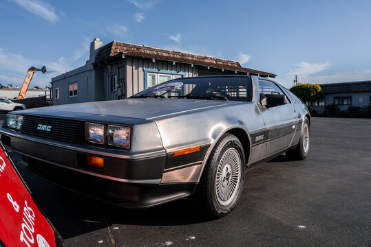 A stainless steel DeLorean DMC 12 sits on a paved lot in Monterey, California. Late afternoon light casts long shadows, showing gullwing seams, wheels, and DMC badge.
