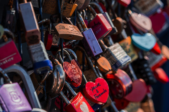 A dense cluster of love locks hangs on a fence in San Francisco, with red and pink heart padlocks, etched initials, warm late afternoon light, bokeh, and worn metal. - Powered by Adobe