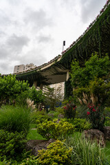 A view of a curved elevated highway completely covered in lush green ivy and climbing plants. The foreground garden contrasts the powerful urban structure with nature's resilience