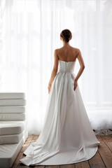 Fototapeta premium Elegant bride in minimalistic white satin wedding gown posing in soft natural light indoors
