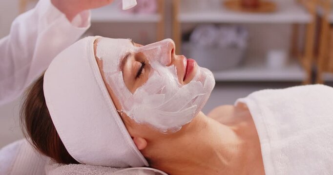 Beautician applying anti-aging facial cream on woman client face to prevent wrinkles in spa salon. Skin care, cosmetic procedures for facial care and beauty treatment concept. 4k video.