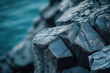 A close-up of dark blue-grey, rugged basalt rock formations with visible cracks and texture, set against a blurred teal ocean or water background.