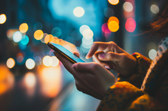 Close-up of woman’s hands using a smartphone in an urban street at night, surrounded by colorful city bokeh lights. - Powered by Adobe