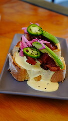 Tostada gourmet con pan artesanal, bañada en cremosa salsa con huevo y coronada con tiras de tocino crujiente, aguacate fresco y cebolla morada encurtida.
