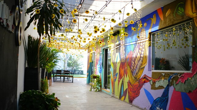 Pasillo decorado con girasoles colgantes y luces c&aacute;lidas, creando un ambiente alegre y acogedor. Al fondo se observan murales coloridos y vegetaci&oacute;n