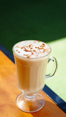 Taza de café con espuma y un toque de canela servida en vaso de cristal, colocada sobre una mesa de madera iluminada por la luz natural.