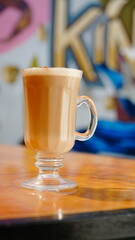 Taza de café con espuma y un toque de canela servida en vaso de cristal, colocada sobre una mesa de madera iluminada por la luz natural.