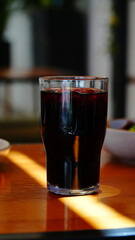 Vaso con bebida oscura servido sobre mesa de madera iluminada por el sol, con fondo desenfocado que resalta el contraste de colores.