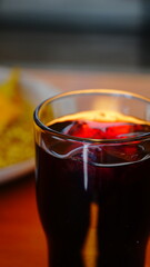Vaso con bebida oscura servido sobre mesa de madera iluminada por el sol, con fondo desenfocado que resalta el contraste de colores.