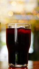 Vaso con bebida oscura servido sobre mesa de madera iluminada por el sol, con fondo desenfocado que resalta el contraste de colores.