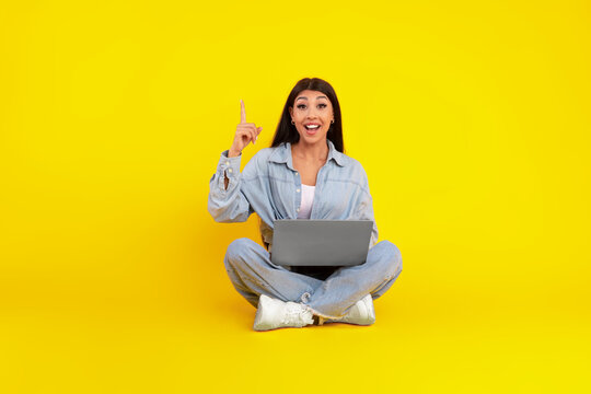 A woman sits cross-legged on a bright yellow floor while holding a laptop. She has a big smile and raises one finger, looking excited as if she just had a great idea.