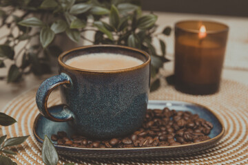 A cup of coffee with milk foam on a blue saucer filled with coffee beans, next to green leaves on a light wooden background. Vintage style, warm tones, and a morning atmosphere.