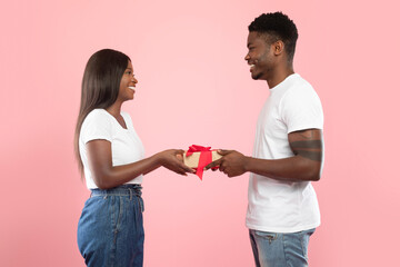 A joyful black couple exchanges gifts in a pink studio. The man presents a gift to his excited partner, celebrating love and togetherness. This moment captures their happiness and affection.