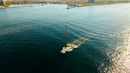 Aerial view Sailboat With Turkish Flag Cruising The Open Sea Under Clear Sky. A lone sailboat glides across calm blue waters, its bright Turkish flag fluttering from the mast. The scene conveys travel