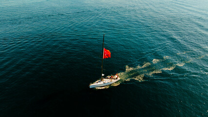 Aerial view Sailboat With Turkish Flag Cruising The Open Sea Under Clear Sky. A lone sailboat glides across calm blue waters, its bright Turkish flag fluttering from the mast. The scene conveys travel