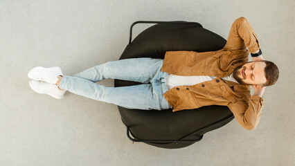 A young man is reclining on a beanbag chair in a cozy indoor setting. He appears relaxed, with his...