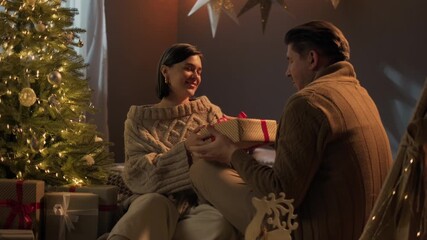 Happy woman is making Christmas gift to his boyfriend at home. Cheerful couple embracing kissing sitting near Christmas tree. Romance, love, holiday and surprise - Powered by Adobe