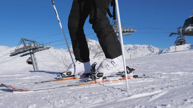 Close up view shows the skier feet in boots, stepping on ski fasteners to fix them before starting skiing on snow covered slope in mountain. Against of a chairlift and Alpine peaks on a sunny day