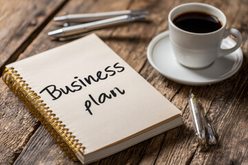 Creative workspace setup featuring a handwritten business plan on a spiral notebook, accompanied by a cup of black coffee and elegant pens on a rustic wooden table