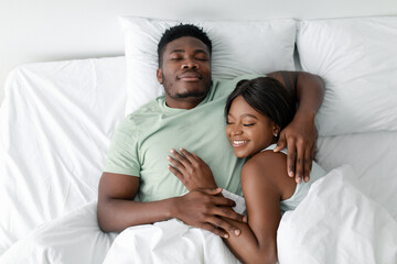 A young couple relaxes in bed, wrapped in each other's arms. They appear content and happy, enjoying a tranquil morning together in soft, white bedding.