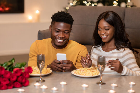Couple celebrates Valentine's Day with a candlelit dinner at home. The husband happily uses his smartphone while his wife enjoys the meal. Roses and soft lighting create a warm atmosphere.