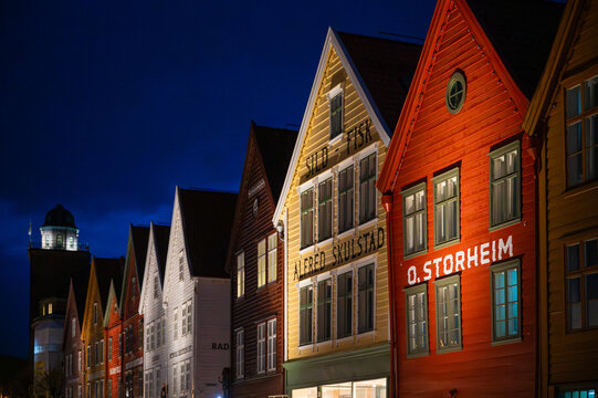 Bryggen, Bergen, Norway, row of colorful, historic buildings illuminated at night, including Sild Fisk and Alered Skulstad, with a tower in the background