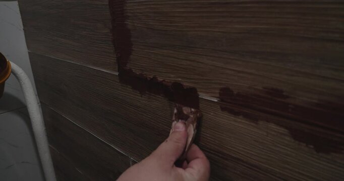 Tiler's hand applying brown grout mixture between wood effect ceramic floor tiles using a small squeegee during a home renovation project, meticulously sealing the joints and seams.