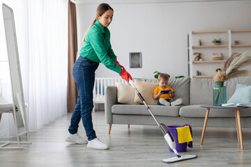 A mother in green cleaning clothes uses a floor mop to tidy the living room. Her young child sits on the couch, focused on a device. The space is bright and inviting.