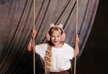Portrait of a girl with long hair wearing pink earmuffs sitting on swing indoors.