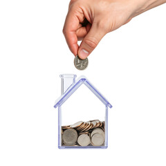 A hand places a coin into a transparent house-shaped piggy bank filled with coins. The background is transparent, emphasizing the savings concept.
