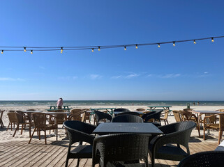 Outdoor beach café with wicker chairs and tables on wooden deck, overlooking calm sea under clear blue sky with string lights above.