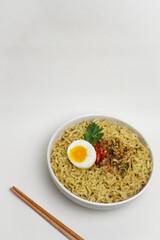 A bowl of instant noodles with half boiled egg, chili, minced meat, and parsley on white background. Copy space for text, minimalist food concept.