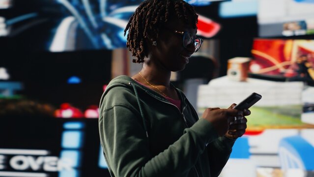 Cheerful young woman using mobile phone to browse internet, looking at surrounded by videos. Smiling african amercian teenager enjoying free time browsing online clips on smartphone, camera B