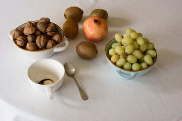 Bodegón con uvas verdes en un bol, granada,  kiwis y nueces en un cuenco, junto a una taza con posos de café y cuchara pequeña.,recibiendo luz suave lateral  sobre fondo claro y neutro.